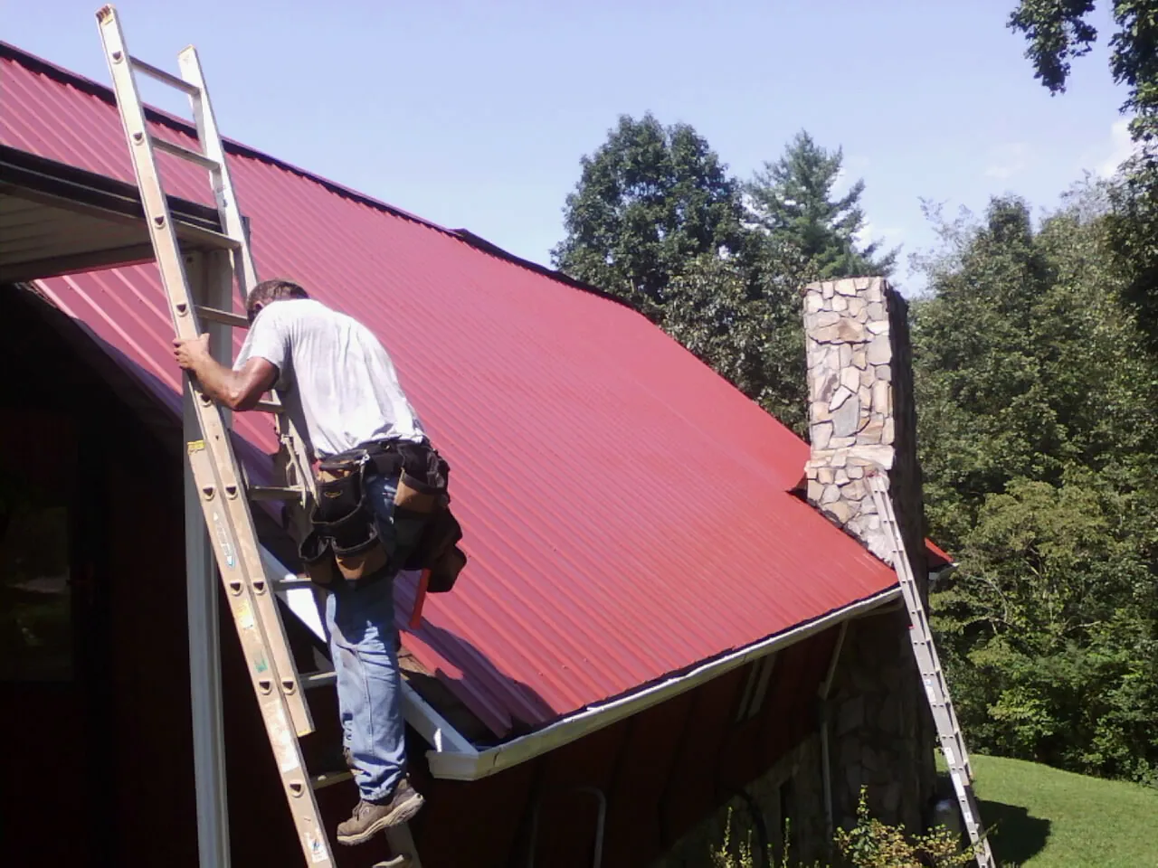 Roofer on a ladder beside a red metal roof and stone chimney — Soffit & Fascia Repair by Roofing Brothers in Needham