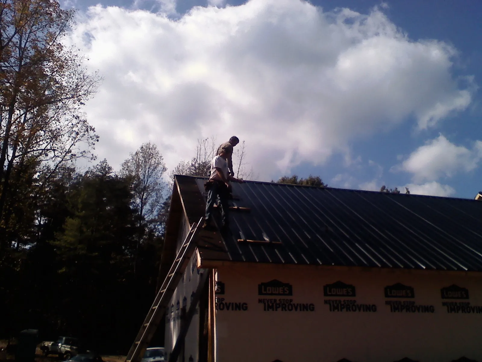 Roofer fastening dark metal panels high against a cloudy sky, from a Roof Inspection job in Needham